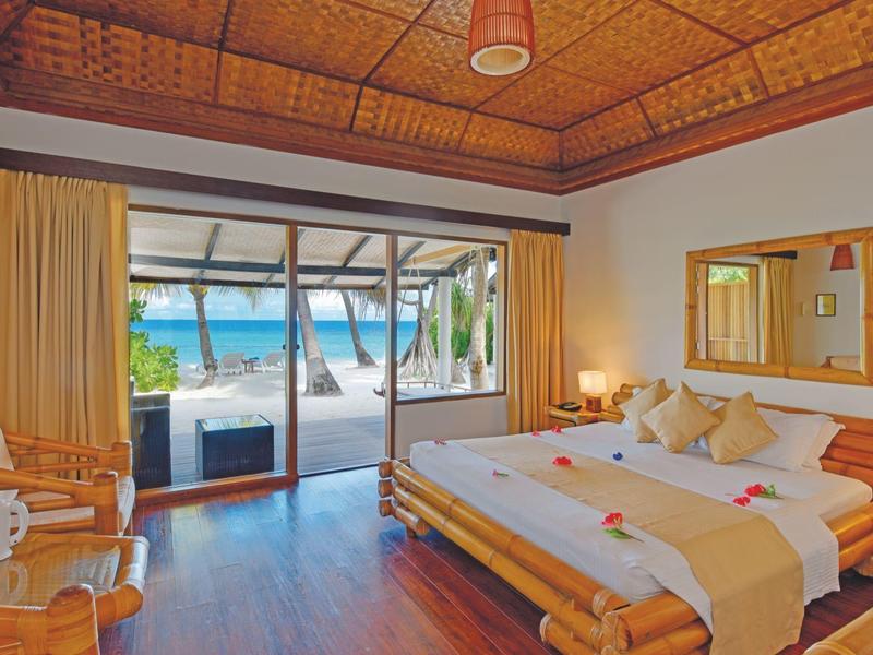 Helles Schlafzimmer mit Holzboden, großem Bett, Stühlen, Blick auf Strand und Meer durch Glasschiebetür.