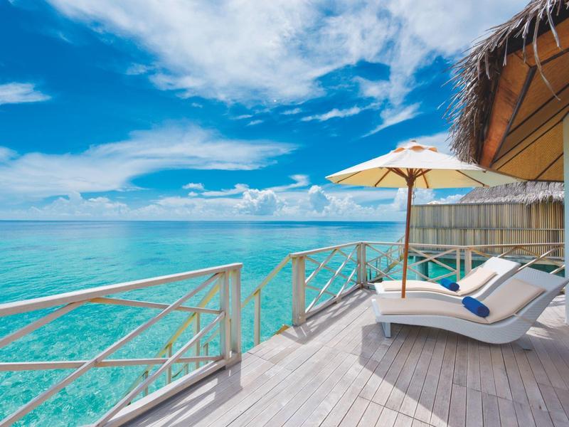 Hölzerne Terrasse mit Liegestühlen und Sonnenschirm am klaren türkisfarbenen Meer unter blauem Himmel.