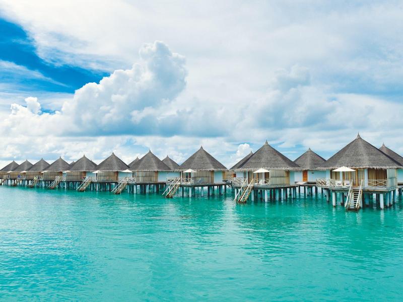 Reihe von Stelzenhäusern mit spitzen Dächern über klarem türkisfarbenem Wasser unter blauem Himmel.