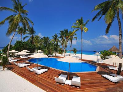 Pool mit Holzdeck und Liegen, umgeben von Palmen und weißem Sandstrand unter blauem Himmel.