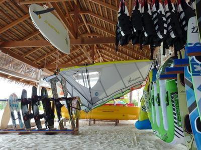 Strandverleih mit Surfbrettern, Segelboot und Neoprenanzügen unter einem Strohdach.