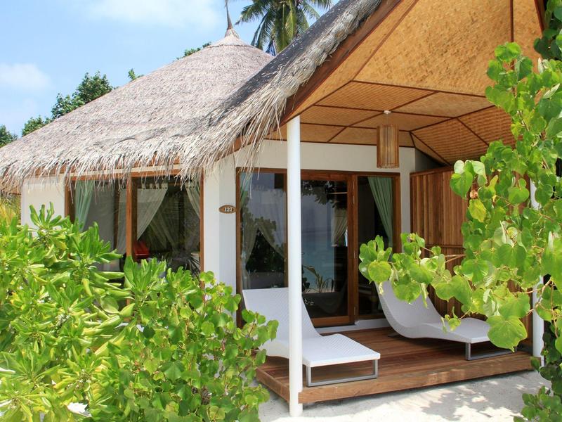Strandbungalow mit Strohdach, Holzveranda und weißen Liegestühlen, umgeben von grünen Pflanzen.
