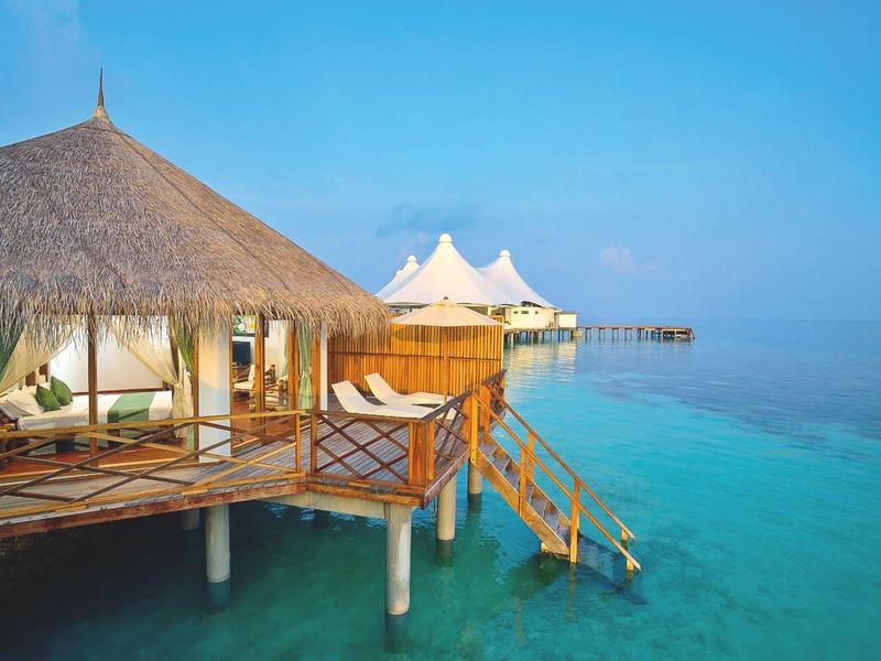 Bungalows auf Stelzen über klarem blauem Wasser mit Treppe ins Meer bei hellem Himmel.