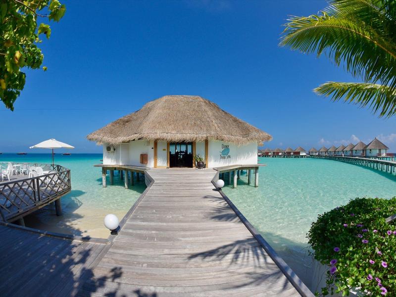 Holzsteg führt zu einem Bungalow über kristallklarem Wasser unter blauem Himmel.