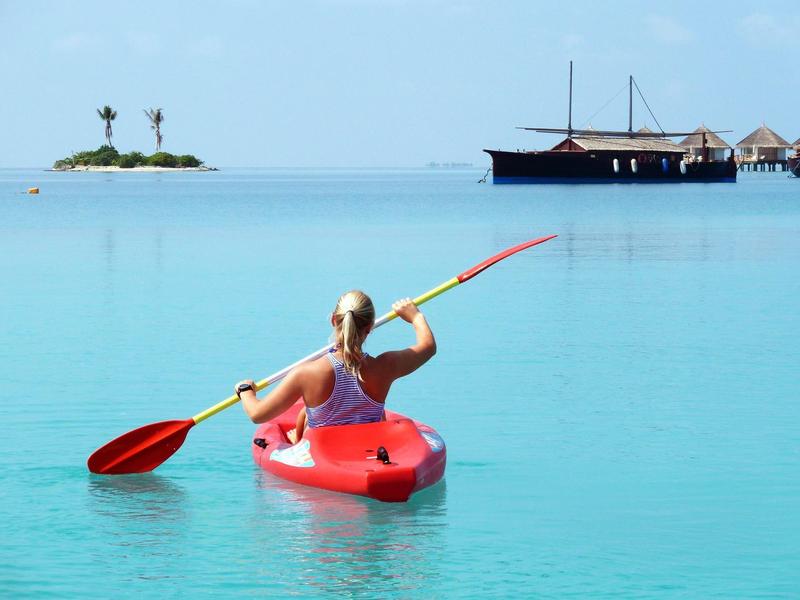 Frau paddelt mit rotem Kajak auf klarem, ruhigem Wasser mit Insel und Boot im Hintergrund
