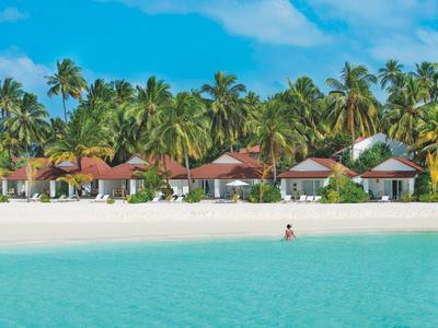 Strand mit weißem Sand, türkisfarbenem Meer, Palmen und kleinen Bungalows im Hintergrund.