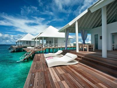 Luxuriöse Strandbungalows mit Holzdielen, weißen Liegen und türkisfarbenem Meer unter blauem Himmel.