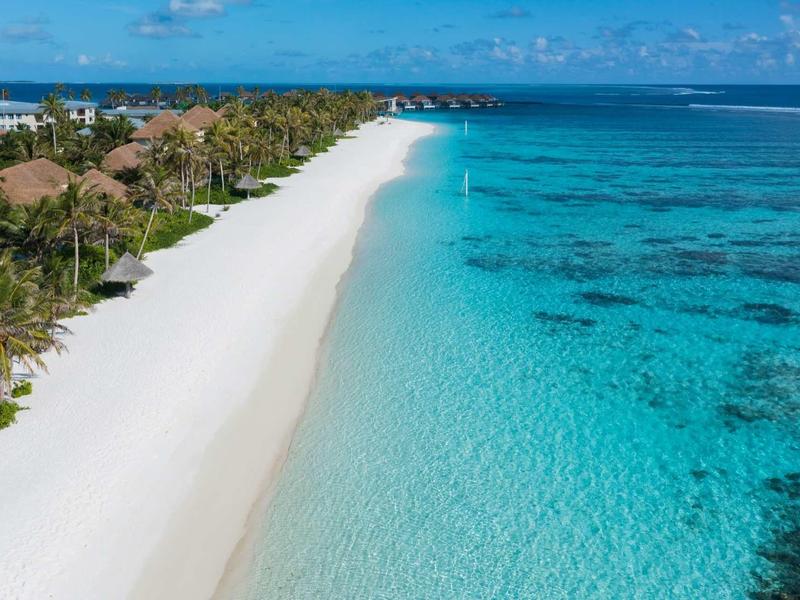 Weißer Sandstrand mit Palmen und Bungalows neben klarem, türkisblauem Wasser unter blauem Himmel.