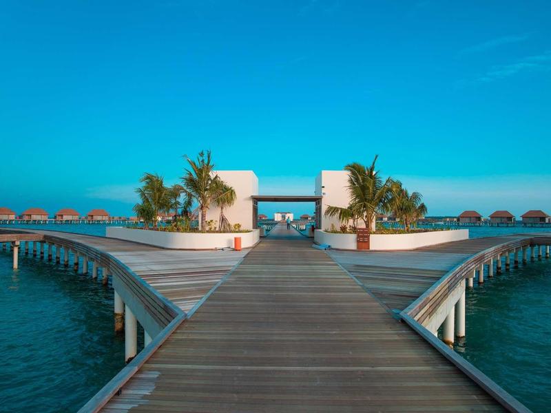 Holzsteg teilt sich zu weißen Bungalows auf ruhigem blauen Wasser mit Palmen und klarer Himmelskulisse.