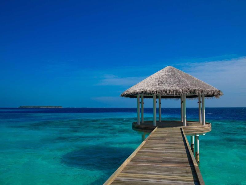 Ein Holzsteg führt zu einer offenen Hütte mit Strohdach über kristallklarem, blauem Wasser.