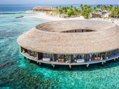 Kruhový plážový resort se střechou pokrytou došky nad čistou tyrkysovou vodou a bílou písečnou pláží.