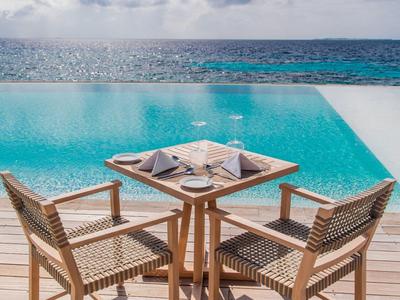 Dřevěný stůl se židlemi na terase u infinity bazénu s výhledem na moře pod modrou oblohou.