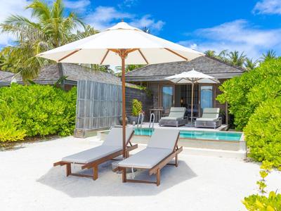 Dvě lehátka pod slunečníkem na bílém písku před bungalovem a modrou oblohou.