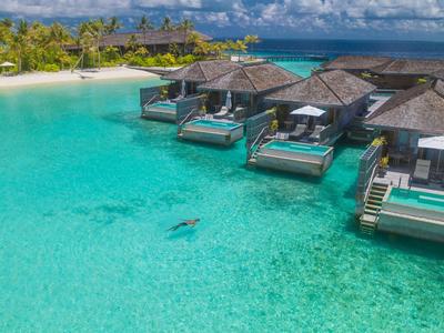 Luxusní bungalovy nad vodou s přímým přístupem ke křišťálově čisté tyrkysové vodě a písečnou pláží v pozadí.