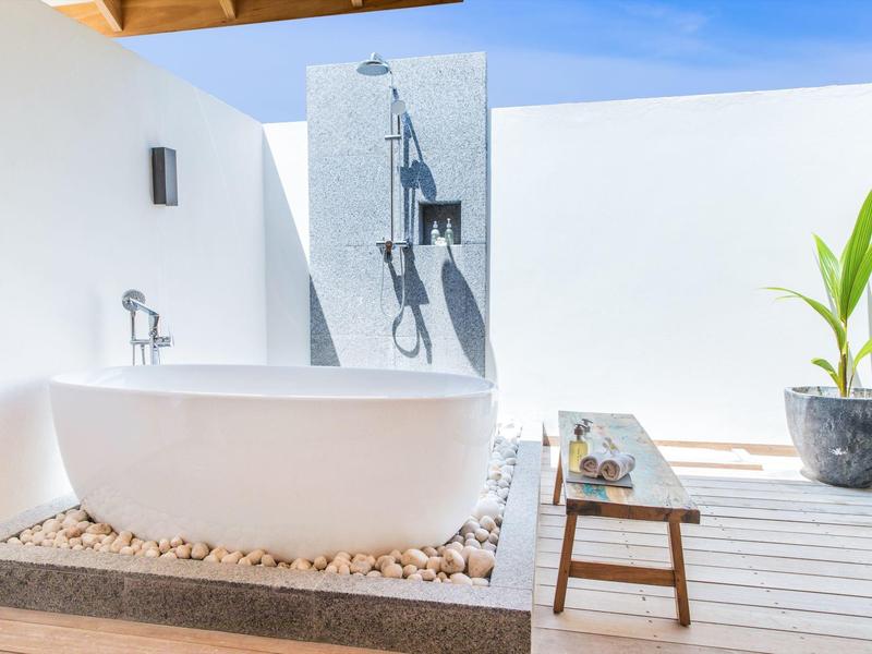 Volně stojící vana na štěrku vedle venkovní sprchy v moderní, světlé venkovní oblasti.