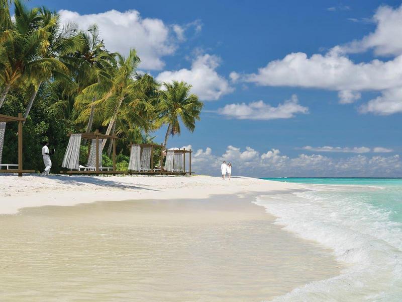 Bílá písečná pláž s palmami a modrou oblohou nad průzračnou tyrkysovou vodou.