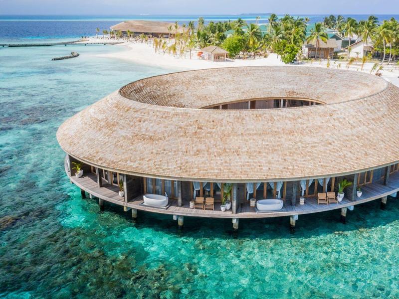 Kruhový plážový resort se střechou pokrytou došky nad čistou tyrkysovou vodou a bílou písečnou pláží.