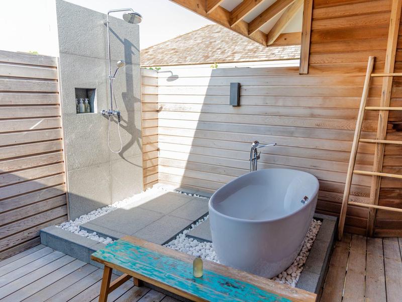Venku koupelna s volně stojící vanou, sprchou a dřevěnými stěnami pod střechou.