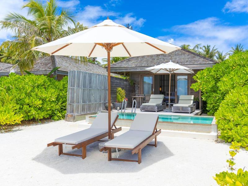 Dvě lehátka pod slunečníkem na bílém písku před bungalovem a modrou oblohou.