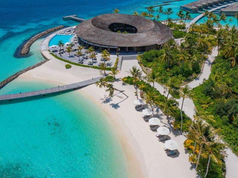 Pláž s čistou tyrkysovou vodou, slunečními deštníky a kulatou budovou s doškovou střechou u moře.