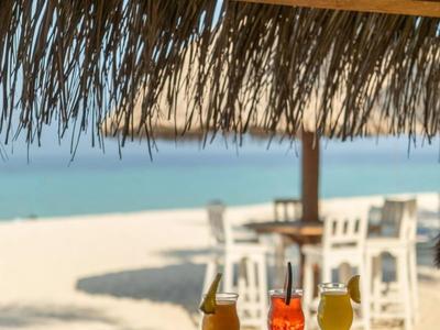 Three colorful cocktails on a counter overlooking a sandy beach and white chairs under a thatched roof.