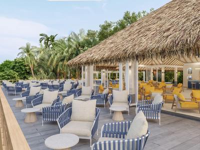 Otwarta przestrzeń wypoczynkowa z krzesłami i stolikami na drewnianym tarasie obok budynku z dachem ze strzechy.