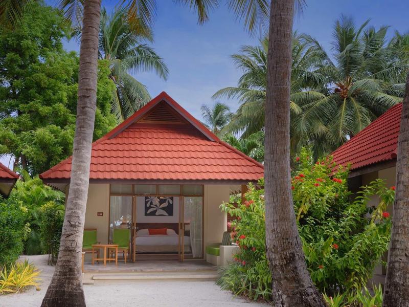 Small bungalow villa with red roof and sun loungers, surrounded by palm trees and tropical vegetation.
