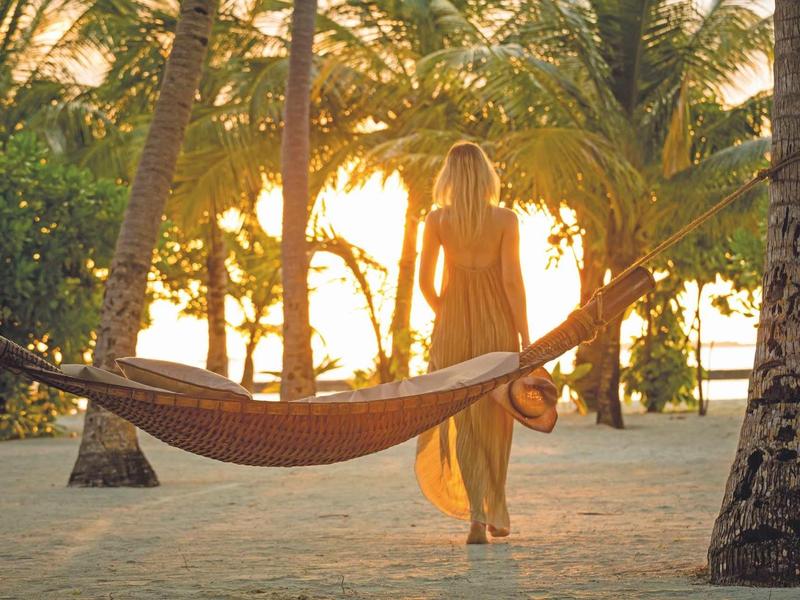 Woman in dress walks at sunset among palm trees on the beach.