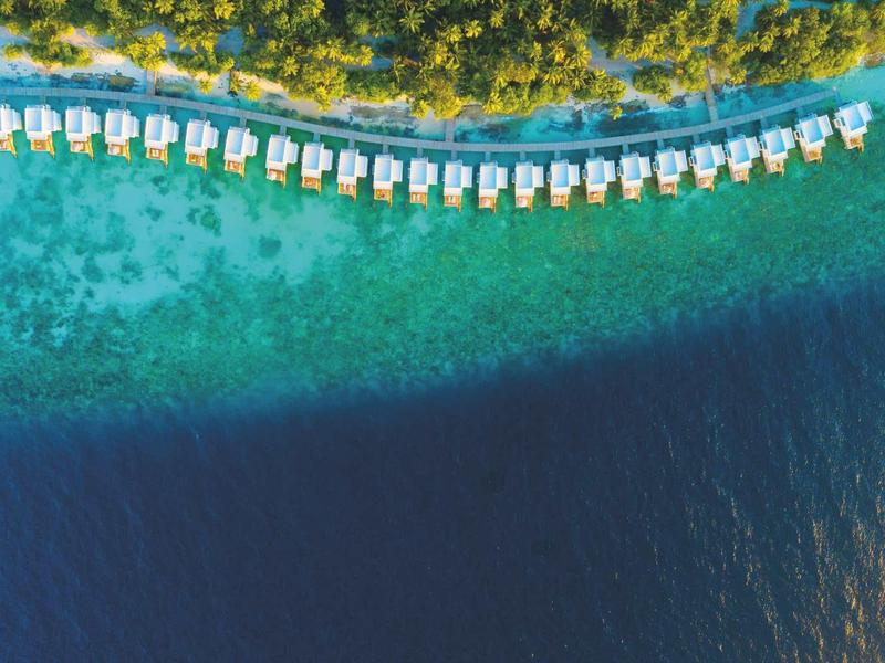 Vue aérienne de bungalows sur l'eau le long d'une forêt verte et d'un océan bleu clair.