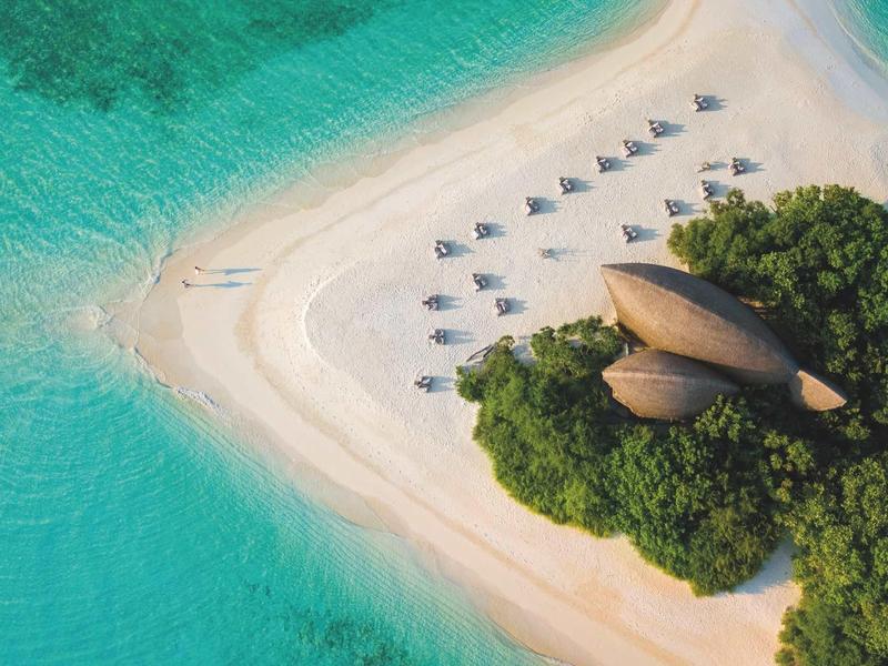 Vue aérienne d'une plage tropicale avec eau turquoise, sable blanc et huttes sous des palmiers.