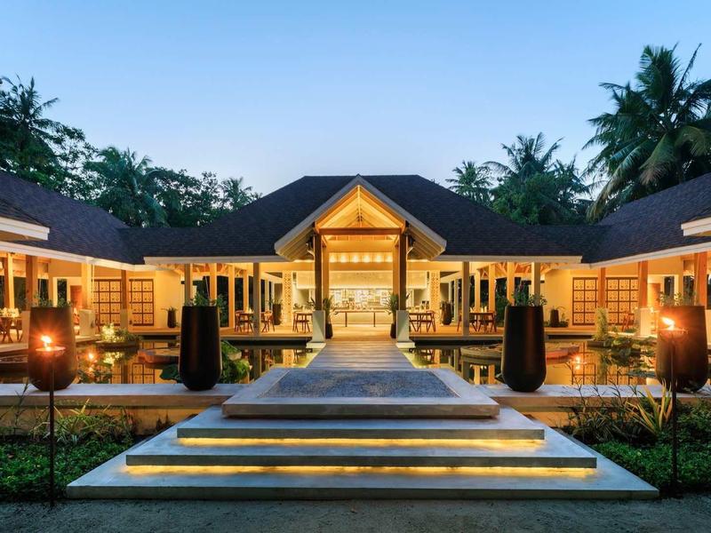 Un hôtel tropical éclairé avec piscine et jardin soigné au coucher du soleil.
