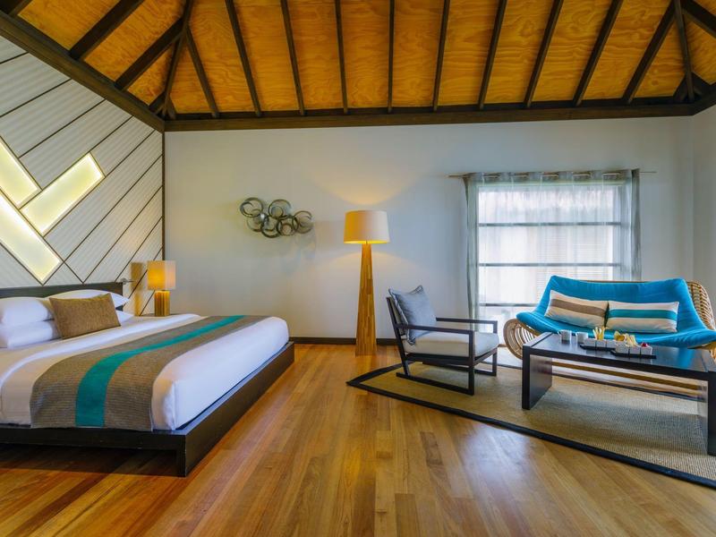 Chambre d'hôtel moderne avec grand lit, sol en bois et coin salon confortable avec canapé et fauteuils.