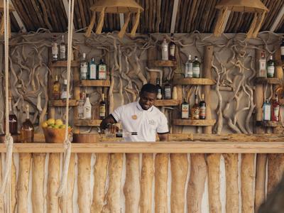 Barkeeper steht hinter Holztresen in rustikalem Strandbar-Umfeld mit Bambusdach.