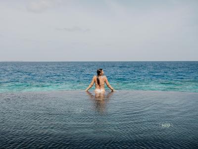 Person sitzt an der Kante eines Infinity-Pools mit Blick auf das offene Meer unter bewölktem Himmel