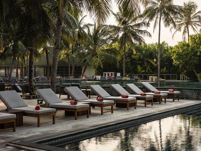 Poolbereich mit Sonnenliegen und roten Handtüchern unter Palmblättern bei Sonnenuntergang.