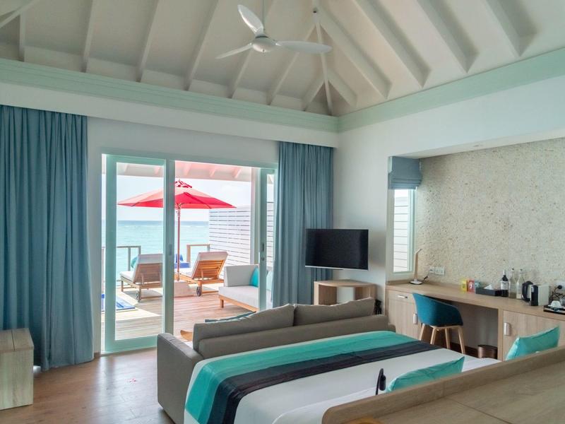 Helles Hotelzimmer mit Meerblick, Bett, Sofa, Schreibtisch und Terrasse mit Loungesessel und Sonnenschirm.