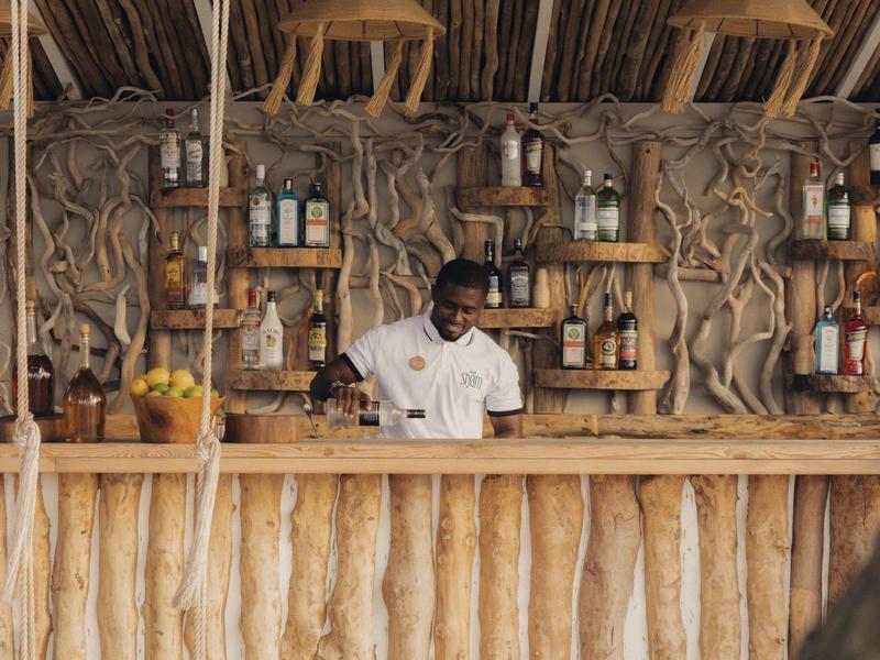 Mann hinter rustikalem Bar mit Holz und Getränken, natürliche Dekoration im Hintergrund.