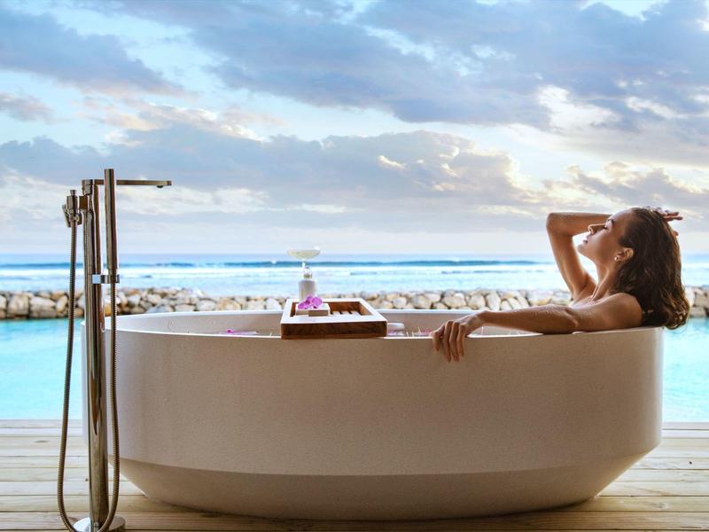Person relaxing in a freestanding bathtub with ocean view and cloudy sky.
