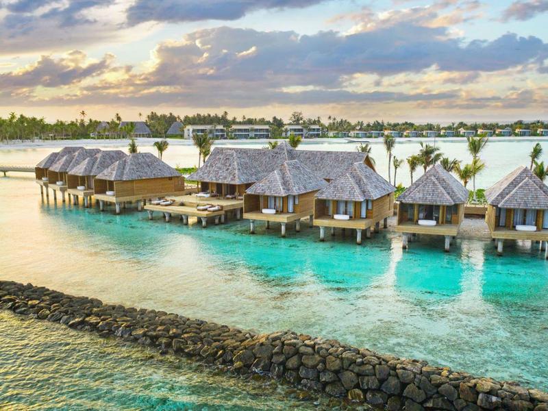 Overwater bungalows at sunset with clear water and palm trees in the background.