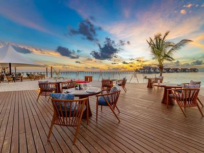 Große Holzterrasse mit Stühlen und Tischen, Palmen, Sonnenuntergang und buntem Himmel am Wasser.