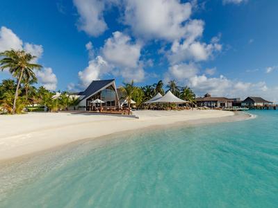 Ein weißer Sandstrand mit türkisblauem Wasser und Palmen vor kleinen Strandbungalows unter blauem Himmel.