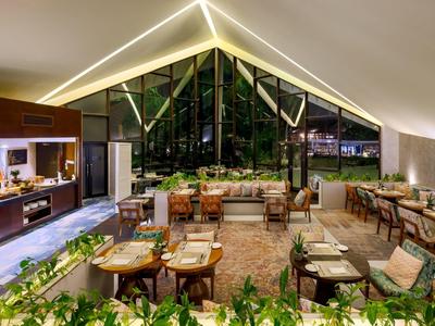 Modernes, gemütliches Restaurant mit viel Holz, großen Fenstern und grünen Pflanzen, warm beleuchtet.
