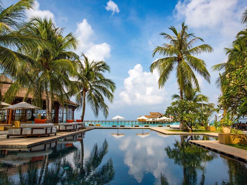 Piscina lujosa con palmeras y vista al mar bajo cielo azul en un resort vacacional.
