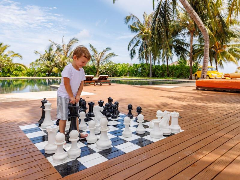 Niño jugando al ajedrez en un tablero grande al aire libre en una terraza de madera con palmeras de fondo.