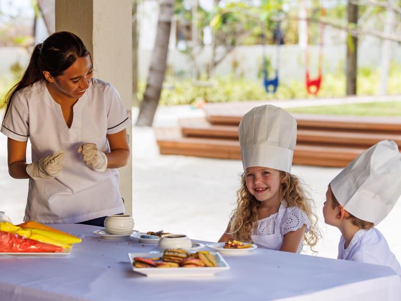 Dos niños con gorros de chef horneando al aire libre, supervisados por una mujer con delantal blanco.