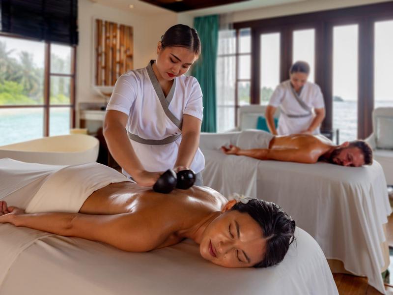 Dos mujeres disfrutan de un masaje relajante en una sala de bienestar luminosa con grandes ventanas.
