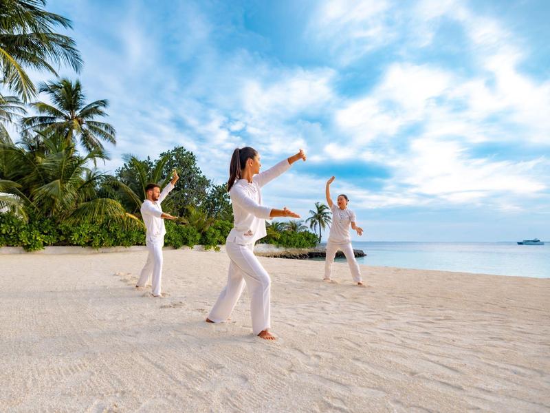 Tres personas practicando tai chi en la playa con palmeras y mar al fondo bajo un cielo nublado