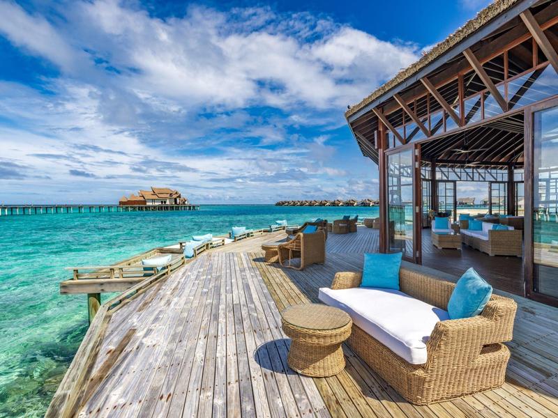 Terraza de madera lujosa sobre agua clara con muebles lounge y vistas al mar con clima soleado.