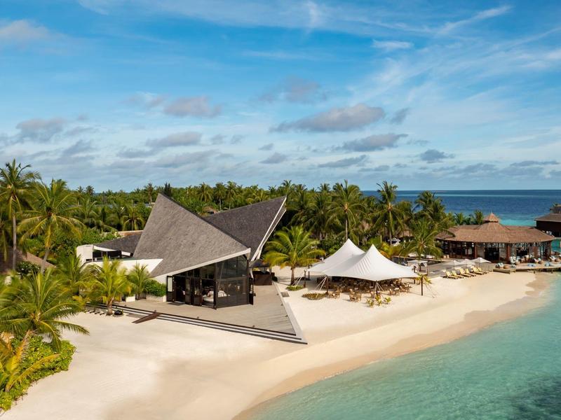 Hotel de playa con arquitectura moderna, palmeras y agua turquesa bajo un cielo despejado.