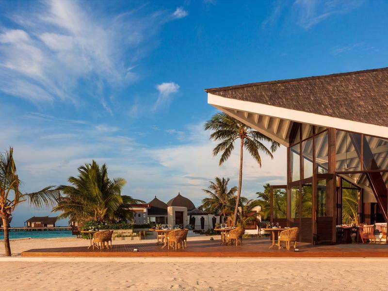 Complejo de resort de lujo en la playa con piscina, palmeras y bungalows frente al mar bajo cielo azul.